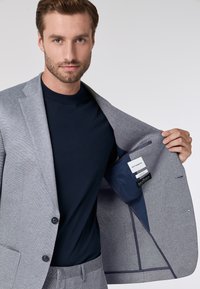 Blazer grigio a trama con due bottoni, indossato su una camicia blu navy. Interno con fodera blu e etichette. Vestibilità su misura e design moderno.