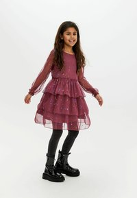 Lila gestufte Kleid mit langen transparenten Ärmeln und funkensprühenden Akzenten, kombiniert mit schwarzen Knöchelstiefeln und glitzernden schwarzen Strumpfhosen.