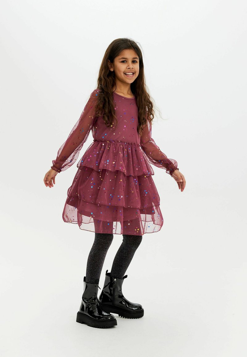 Lila gestufte Kleid mit langen transparenten Ärmeln und funkensprühenden Akzenten, kombiniert mit schwarzen Knöchelstiefeln und glitzernden schwarzen Strumpfhosen.