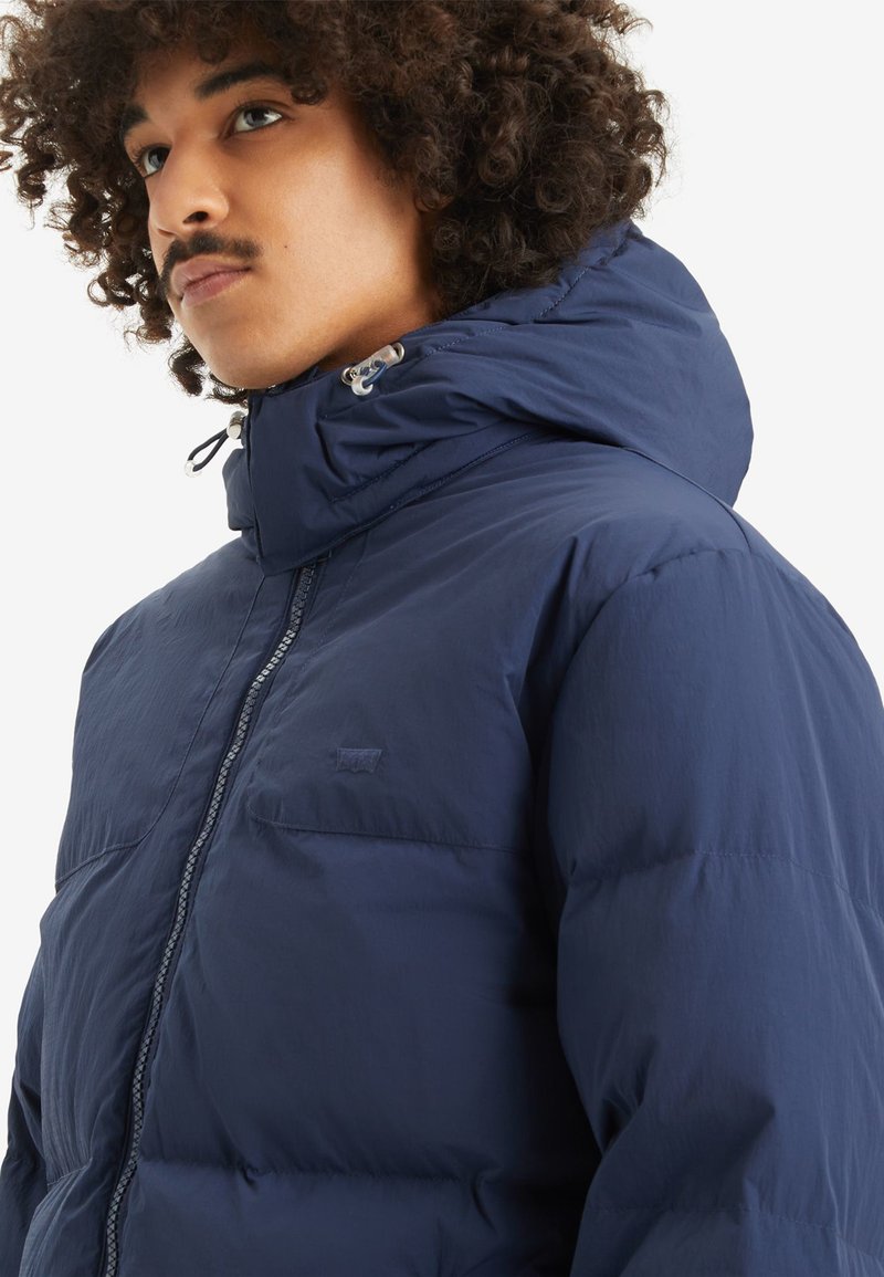 Chaqueta acolchada azul marino con cuello alto, cierre de cremallera y bolsillo frontal. Cuenta con un tejido texturizado y cordones en la capucha.