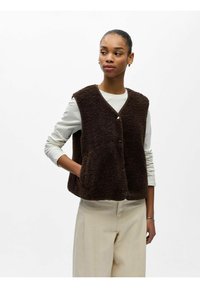 Femme portant un gilet sans manches en peluche marron foncé sur une chemise blanche à manches longues et un pantalon beige, debout avec une main dans la poche.