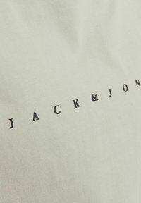 Hellgrünes Baumwoll-T-Shirt mit einem schwarzen Logo-Druck, der "JACK & JON" in einer ordentlichen, weit auseinander stehenden Schriftart zeigt. Glatte Textur.