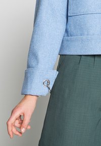 Světle modrá vlněná bunda se strukturovaným límcem, ohrnutými manžetami a stříbrnými detaily na rukávu, spárovaná se zelenými textured kalhotami.