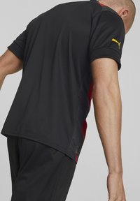 Hombre vistiendo un chándal negro con paneles laterales rojos y un logo amarillo de Puma en la manga, inclinado hacia adelante sobre un fondo gris.