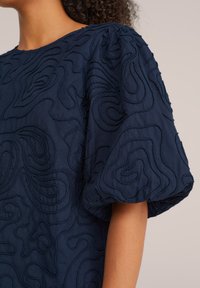 Blusa azul marino con mangas abullonadas, adornada con patrones de remolinos bordados y tejido texturizado para añadir dimensión.