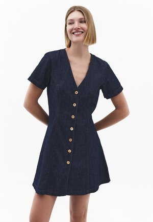 Kvinde med kort blondt hår iført en kortærmet mørk denimkjole med træknapper foran, stående med hænderne bag ryggen.