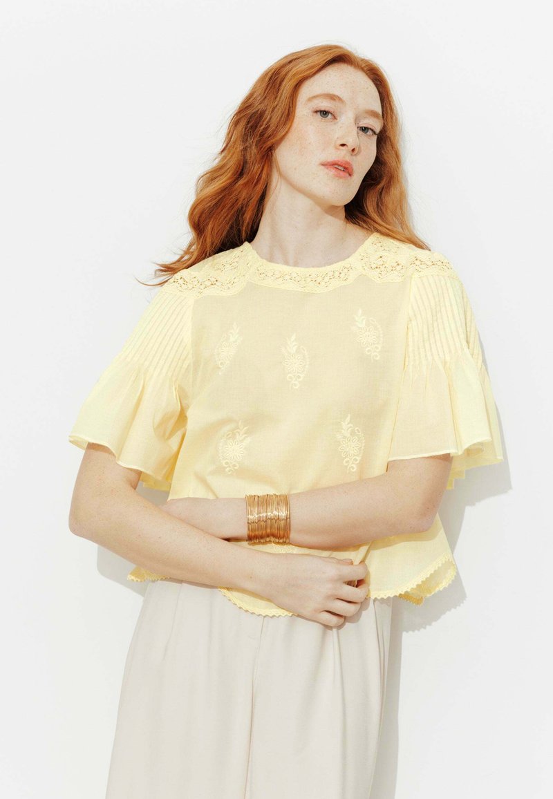 Femme aux cheveux roux portant un chemisier brodé jaune pâle à manches évasées et un pantalon beige clair, les bras croisés avec un bracelet en or.