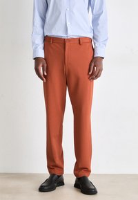 Pantalon texturé orange avec une fermeture à boutons, présentant une coupe droite, associé à une chemise à rayures bleues à boutons et des chaussures noires.