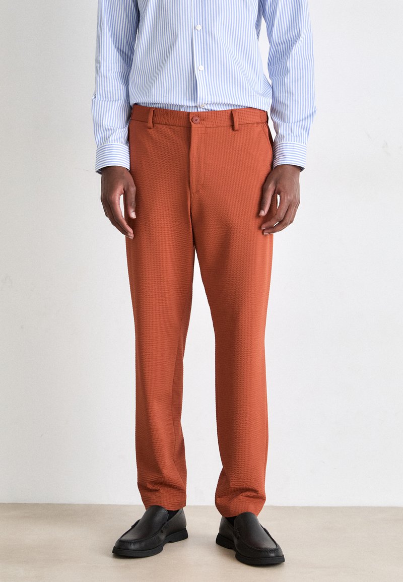 Pantalon texturé orange avec une fermeture à boutons, présentant une coupe droite, associé à une chemise à rayures bleues à boutons et des chaussures noires.