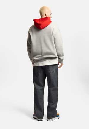 Mężczyzna z ogolonymi włosami, ubrany w szarą bluzę z kapturem w czerwonym kolorze, ciemne dżinsy i kolorowe trampki, stoi tyłem na białym tle.