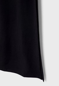 Vêtement en tissu noir avec une texture lisse, présentant une coupe droite et un détail d'ouverture sur les côtés, suspendu devant un fond neutre.