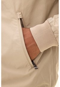 Beige Jacke mit einer Reißverschlusstasche und elastischen Bündchen. Der Stoff wirkt glatt mit matter Oberfläche, Details sind rund um die Tasche sichtbar.