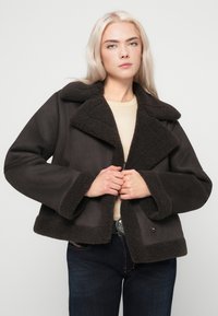 Nuori nainen, jolla on pitkät vaaleat hiukset, pukeutuneena tummanruskeaan lammasnahkatakkiin, beigeen puseroon ja tummiin farkkuihin, yksinkertaista taustaa vasten.