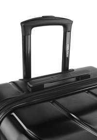 Maleta negra con una superficie texturizada, que cuenta con un asa retráctil y cierre, marcada con "100% DKNY" en el lado del asa.