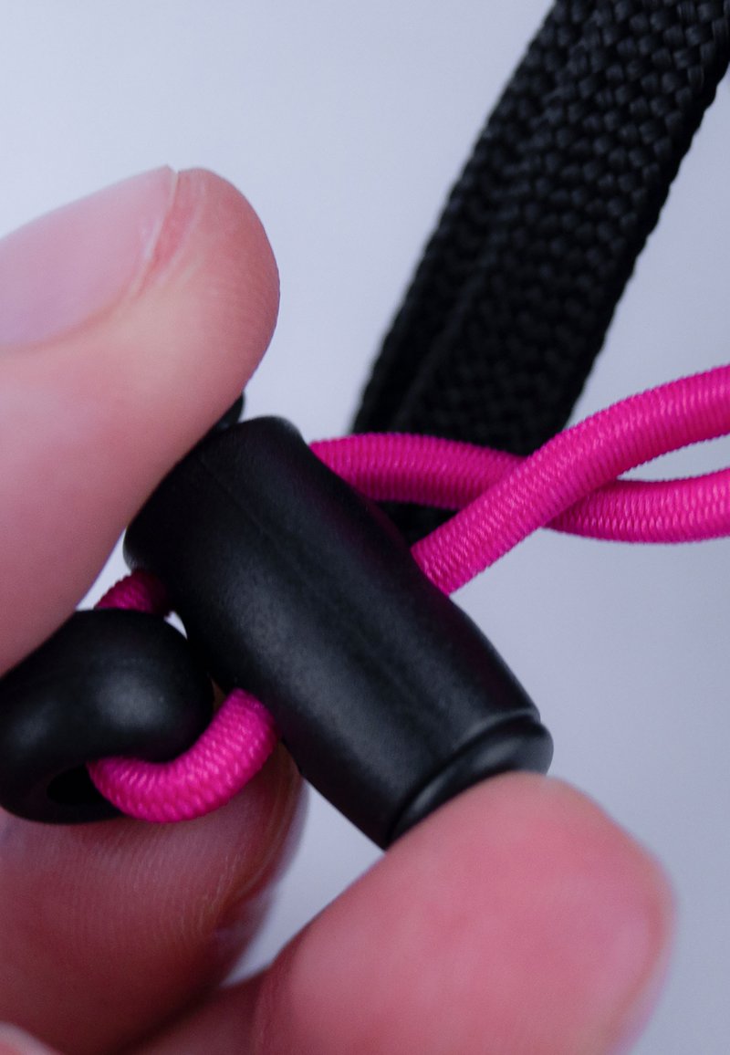 Schwarzer Kordelstopper sichert eine leuchtend pinke Schnur, gehalten zwischen Zeigefinger und Daumen, mit gewebtem Texturdetails an Schnur und Stopper.