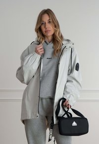Hvid vandtæt jakke med elastiske manchetter, grå hættetrøje, grå sweatpants og en sort taske med et trekantet logo og justerbar rem.