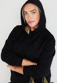 Črna kapuca z teksturiranim čipkastim prelivom na ramenih, z zadnjim žepom in nastavljivimi vrvmi na kapuci.
