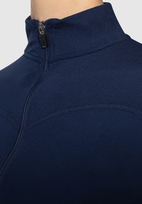 Marinblå zip-up jacka i mjukt, elastiskt tyg. Har en hög krage och synliga sömdetaljer längs axlarna och bröstet.