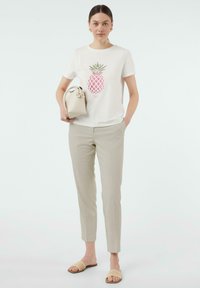 White T-shirt with a pink pineapple graphic, beige tailored pants, and woven tan slide sandals; holding a small cream handbag.