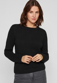 Μαύρο πλεκτό πουλόβερ με υφασμάτινη επιφάνεια, στρογγυλή λαιμόκοψη και ριγωτούς μανικέτες. Φορεμένο πάνω από γκρι παντελόνι, αναδεικνύοντας μια άνετη εφαρμογή.