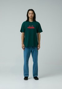 Green short-sleeve T-shirt with red "FORMER" graphic, paired with blue jeans and black shoes. Simple, relaxed fit design and smooth texture.