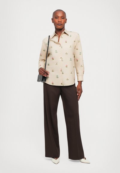 Cămașă button-up bej deschis cu broderie florală, asociată cu pantaloni largi maro închis. Geantă de umăr neagră și pantofi cu vârful ascuțit albi.