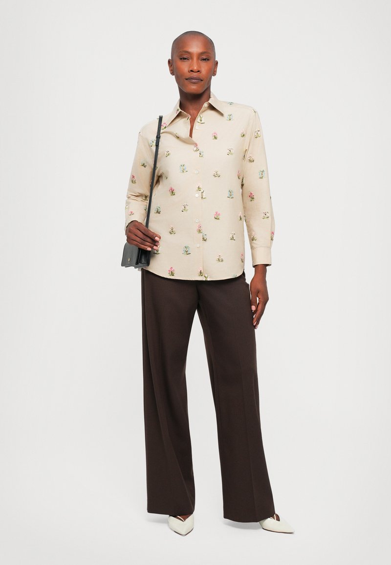 Jasnobeżowa koszula z guzikiem z kwiecistym haftem, połączona z ciemnobrązowymi spodniami z szerokimi nogawkami. Czarna torebka na ramię i białe szpilki z czubkiem.