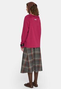 Różowa bluzka z długim rękawem i zaokrąglonym dekoltem, z logo na plecach, połączona z midi spódnicą w kratę w odcieniach szarości, brązu i burgundu.