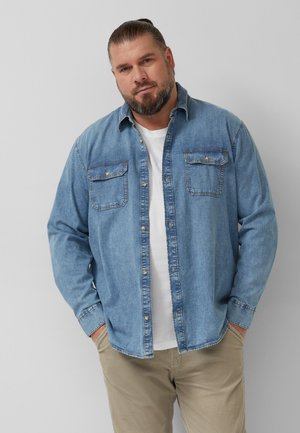 Mann mit Bart und zurückgebundenen Haaren, der ein hellblaues Jeanshemd über einem weißen T-Shirt und beige Hose trägt, steht mit den Händen in den Taschen.