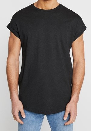 Muž v jednoduchém černém tričku s vyhrnutými rukávy a světlých modrých džínách, stojící s uvolněnými rukama podél těla.