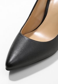 Chaussure à talon haut noire en matériau synthétique texturé, pointe effilée et intérieur lisse de couleur beige. Conçue avec une silhouette élégante.