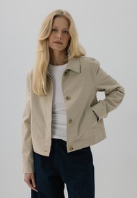 Beżowa kurtka z kołnierzykiem, z przodem zapinanym na guziki i bocznymi kieszeniami, w połączeniu z białą bluzką i ciemnoniebieskimi spodniami. Gładka tekstura.