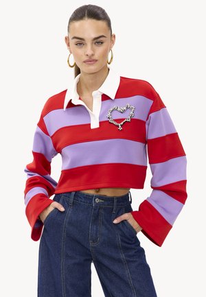 Polo court rayé rouge et violet clair avec un col blanc et un ornement en forme de cœur sur la poitrine. Manches cloche et texture douce.
