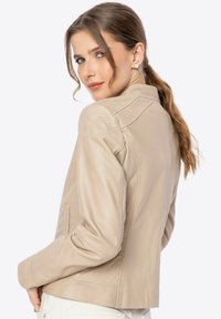 Beige leren jas met een aansluitend ontwerp, zichtbare stiksels en een gladde textuur. Heeft een kraag en zijzakken.