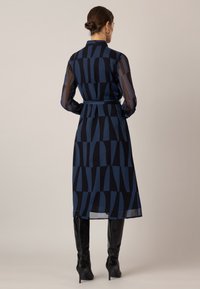 Long navy dress featuring a geometric pattern in varying shades of blue. Made of sheer material, with a tied waist and long sleeves.