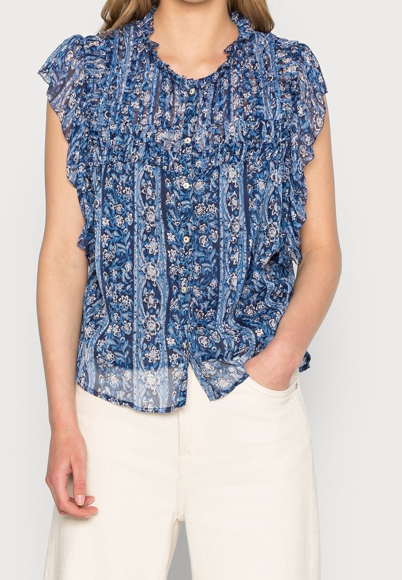 Femme portant un chemisier bleu à fleurs sans manches avec des volants aux épaules et un pantalon blanc, une main dans la poche.