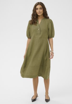 Femme aux cheveux bouclés portant une robe longueur genou vert olive avec des manches bouffantes, un col boutonné, et des escarpins noirs à bride arrière.