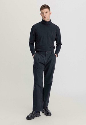 Maglione a collo alto nero e pantaloni scuri, con fronte plissettato, tasche laterali e scarpe nere con suola spessa.
