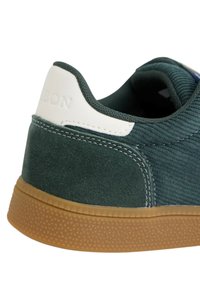 Zapatilla de ante verde con textura acanalada, acento en blanco en el talón y suela de goma con un diseño de banda de rodadura patrón.