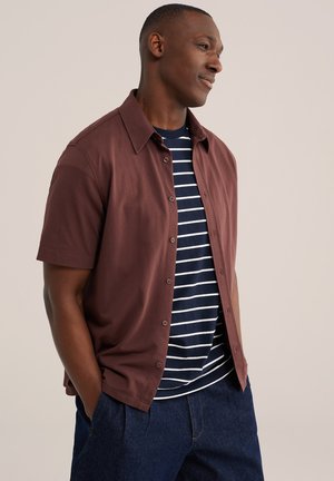 Homme portant une chemise marron à manches courtes boutonnée sur un t-shirt rayé bleu marine et blanc, les mains dans les poches d'un pantalon bleu foncé, faisant face à droite.