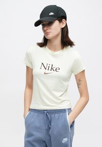 Νέα γυναίκα με καφέ μαλλιά, φορώντας μαύρο καπέλο Nike, κρέμ t-shirt Nike και μπλε παντελόνια Nike, χέρια στις τσέπες, ουδέτερη έκφραση.