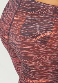 Tights med en mörk bas som har ett vågigt mönster av orange prickar och linjer. Materialet verkar elastiskt med tydliga sömmar.