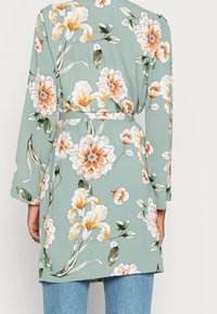 Kimono floral en tejido verde suave con flores naranjas y blancas. Tiene mangas largas, cinturón de lazo y un corte holgado que llega hasta la mitad del muslo.