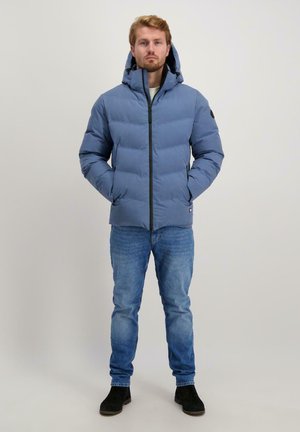Lichtblauwe pufferjack met een voorkantrits, capuchon en zwarte accenten, gecombineerd met een blauwe spijkerbroek en zwarte laarzen. Geplaatst tegen een neutrale achtergrond.