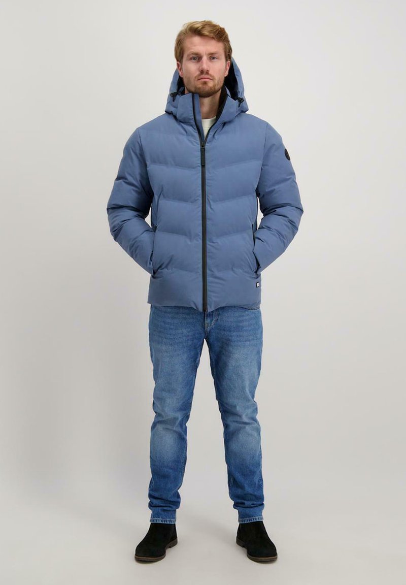 Lichtblauwe pufferjack met een voorkantrits, capuchon en zwarte accenten, gecombineerd met een blauwe spijkerbroek en zwarte laarzen. Geplaatst tegen een neutrale achtergrond.