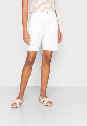 Frau trägt weiße knielange Shorts, beige-weiß gestreiftes ärmelloses Oberteil und weiße Sandalen mit gekreuzten Riemen, steht vor einfarbigem Hintergrund.