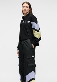 Γυναίκα που φορά μαύρη φόρμα με λωρίδες σε σχήμα βέλους σε ανοιχτό μωβ και ανοιχτό κίτρινο στα μανίκια και στα πόδια, όρθια μπροστά σε απλό φόντο.