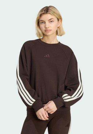 Junge Frau mit blonden Haaren, die einen dunkelbraunen Adidas-Sweatshirt und passende Hose trägt, steht mit verschränkten Händen.
