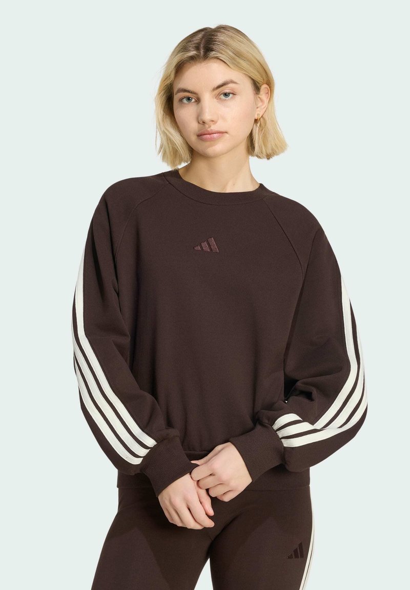Young woman with blonde hair wearing a dark brown Adidas sweatshirt and matching pants, standing with hands clasped.