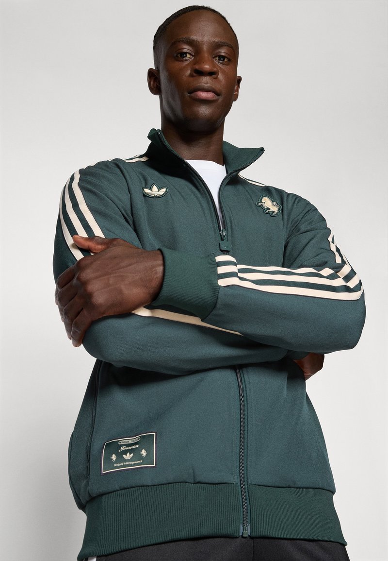 Dunkelgrüne Zip-Jacke mit weißen Streifen, besticktem Logo auf der Brust, gerippten Bündchen und einem Etikett auf der unteren linken Seite. Strukturiertes Gewebe.